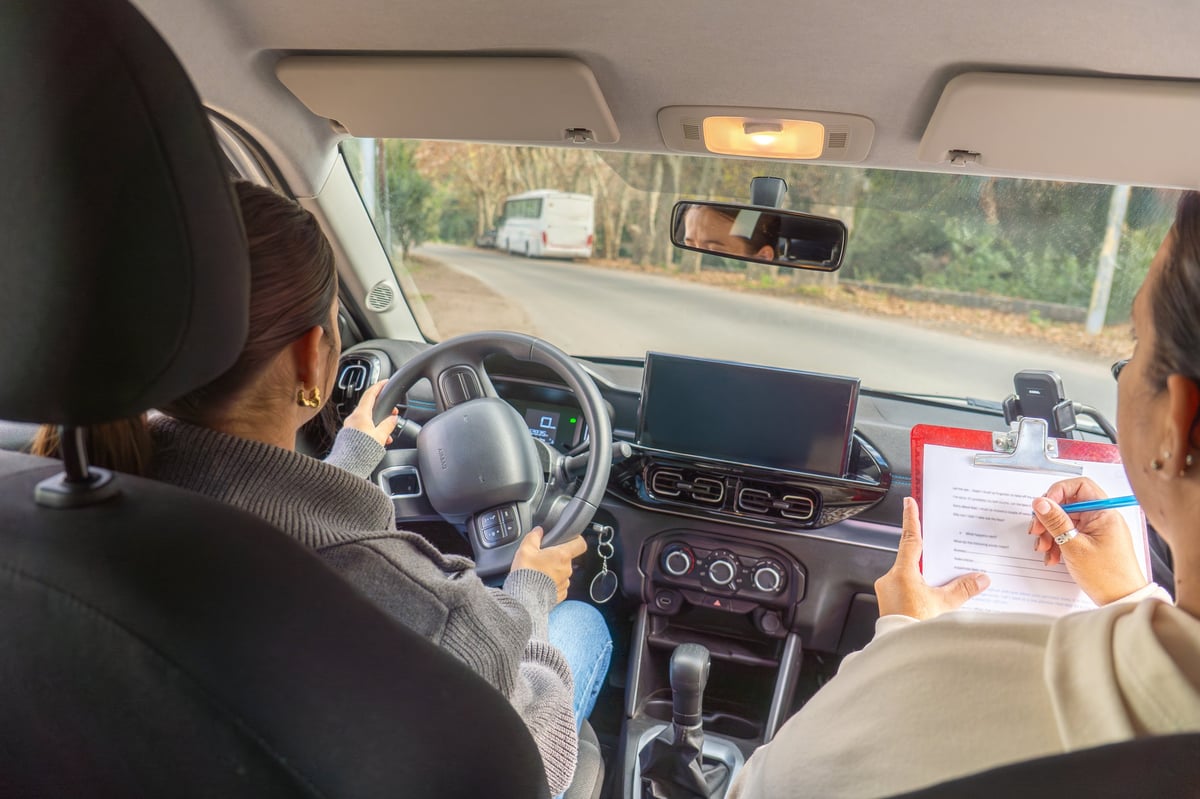 Driving instructor writing on a clipboard while a young woman student drives, dashboard and rearview mirror visible