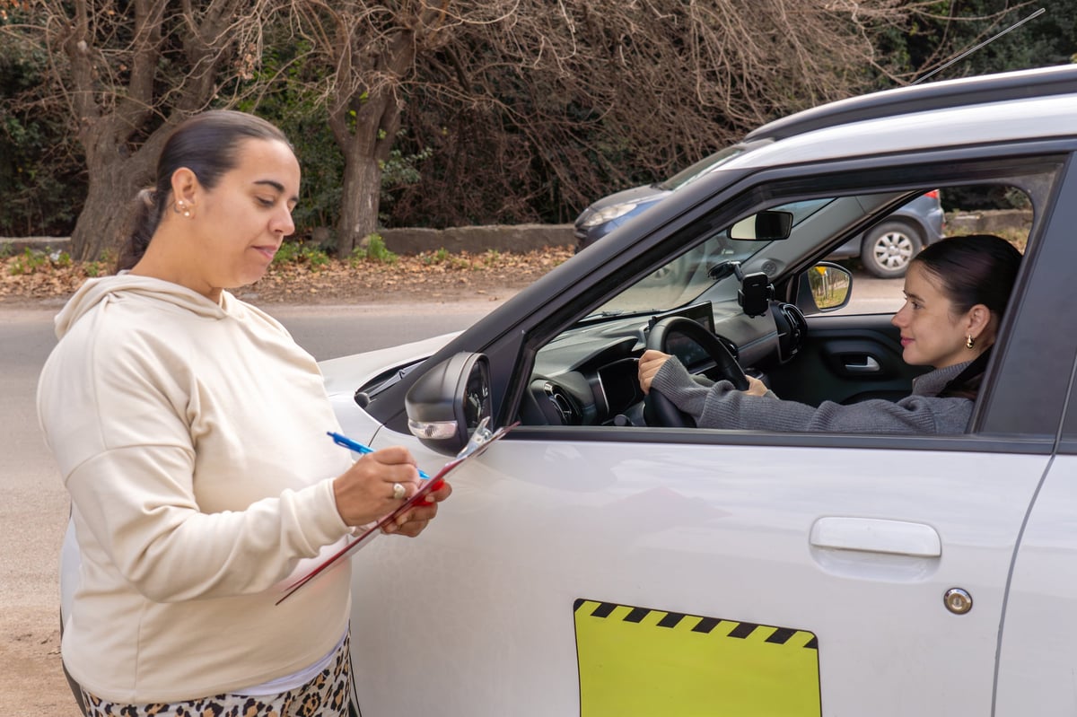 Female driving test examiner writing on clipboard outside car while smiling candidate sits behind the wheel, exam evaluation concept