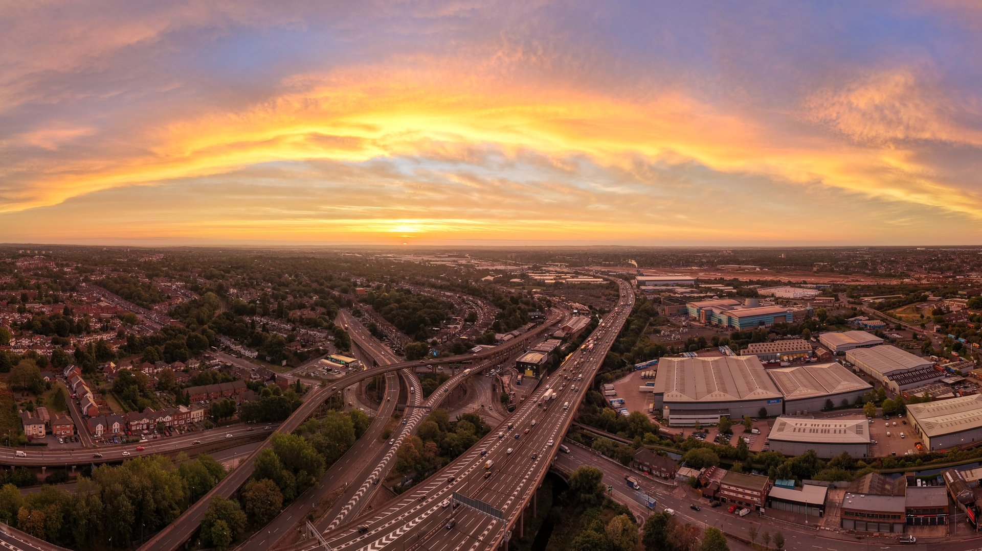 Wide panoramic aerial view of a sprawling motorway interchange at golden sunset, with sweeping roads, busy traffic and surrounding urban neighbourhoods